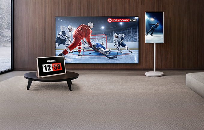 모던한 거실에 ICE HOCKEY LIVE 라고 쓰여진 문구와 아이스 하키 경기 장면의 대형 TV, NEXT GAME 12:58 라고 쓰여진 탁상 시계, 피겨 스케이팅 장면의 스탠바이미가 놓여 있다.